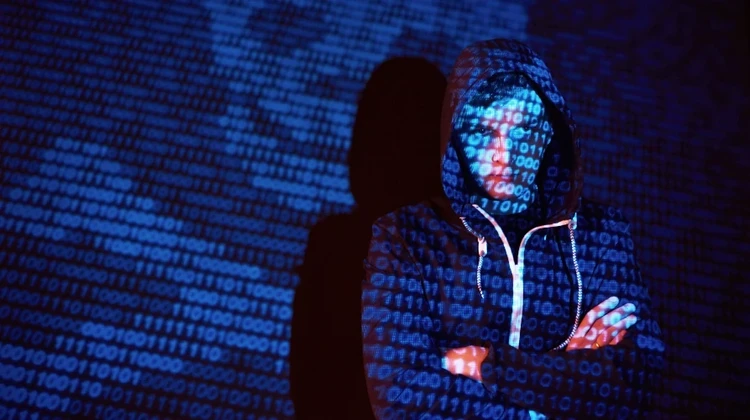 hacker usando moleton com capuz em frente a uma tela de projeção com código binário e ícone de caveira de ossos projetados sobre seu corpo e rosto