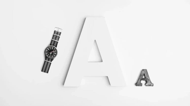 Duas letras A em fontes diferentes em um fundo branco ao lado de um relógio de pulso preto