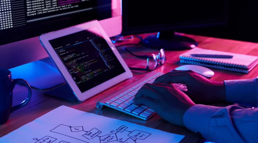 Foto de mãos masculinas digitando num teclado de Mac e um iPad mostrando linhas de código