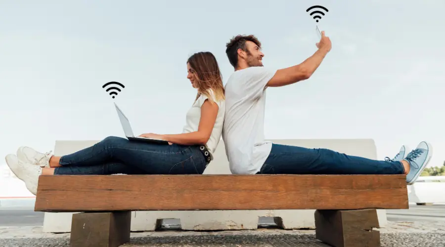 Mulher com notebook e homem com celular, sentados num banco de parque, compartilhando wi-fi