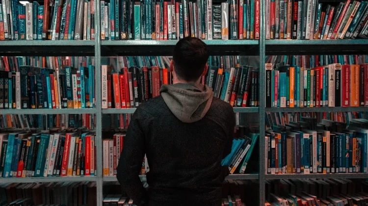 Homem de costas olhando uma estante com prateleiras repletas de livros em uma biblioteca