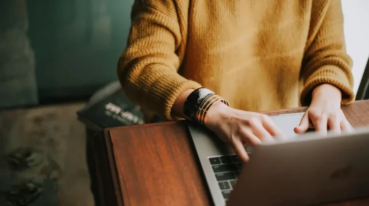 Mulher vestindo suéter marrom digita em um MacBook sobre uma mesa de madeira antiga
