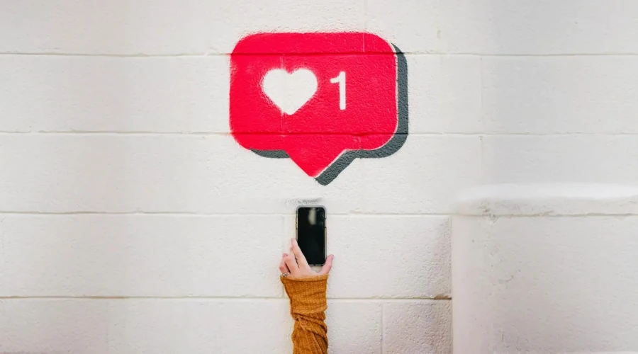 Parede com pichação do ícone de like recebido do instagram com uma mão feminina segurando um smartphone abaixo