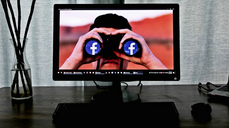 Computador com papel de parede que mostra homem com binóculos apontado na direção da câmera e logos do Facebook no lugar das lentes
