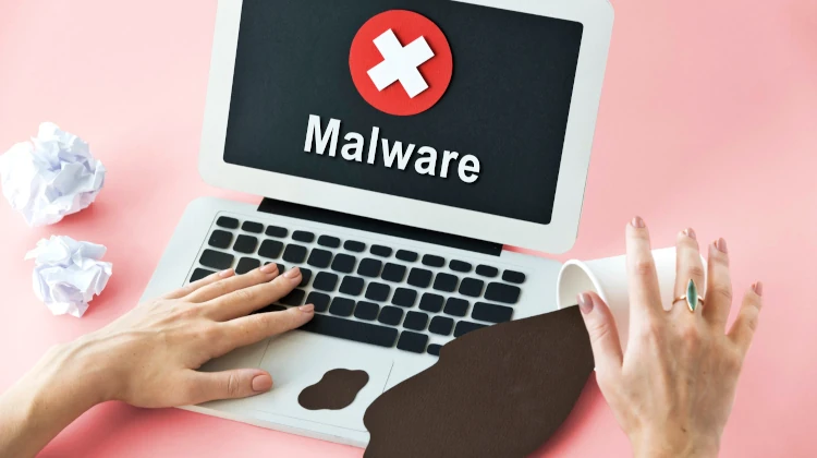 Duas mãos femininas sobre Macbook feito com papel recortado, com palavra MALWARE na tela, ao lado de um copo virado com café derramado