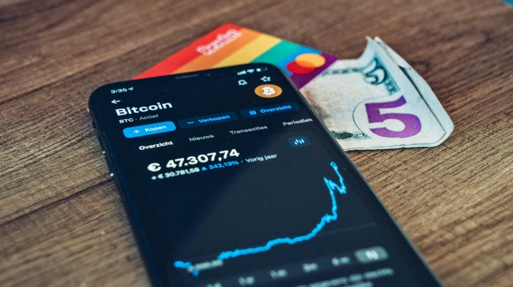 Smartphone sobre uma mesa com cotação do Bitcoin aberta, sobre notas de dinheiro e um cartão de crédito