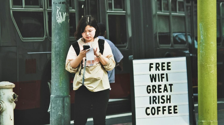 Garota asiática segurando celular ao lado de uma placa escrita free wifi