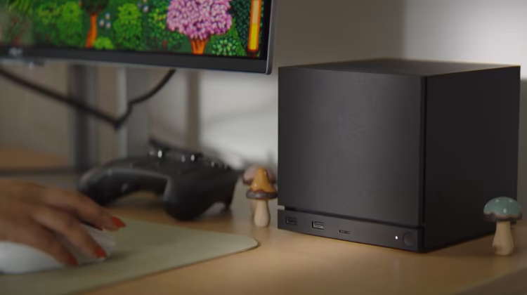 Uma Steam Machine ao lado de um monitor de computador e uma mão feminina segurando um mouse sobre uma mesa de madeira clara