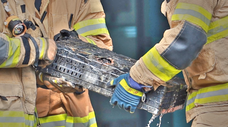 Foto de uma caixa preta queimada e derretida sendo segurada por três bombeiros vestindo roupas de proteção. Não é possível ver seus rostos